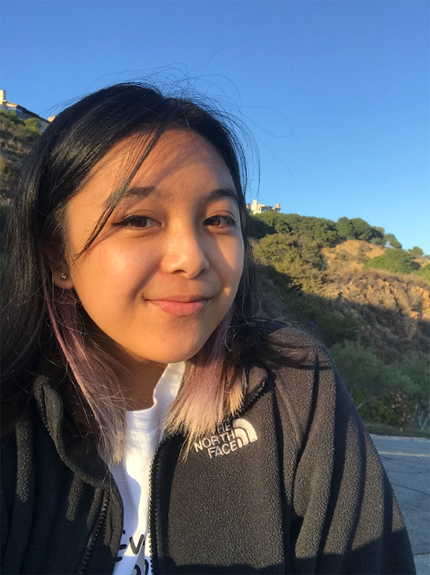 Nuriel is outdoors smiling. There is blue sky and a green hillside behind them.