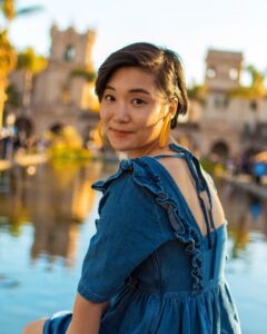 Carolyn is outdoors by a body of water smiling at the camera while looking over their shoulder.