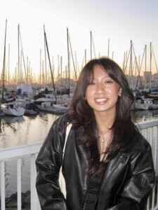 Michelle smiles on a pier, with a sunset and rows of boats behind her. She is wearing a black leather jacket.